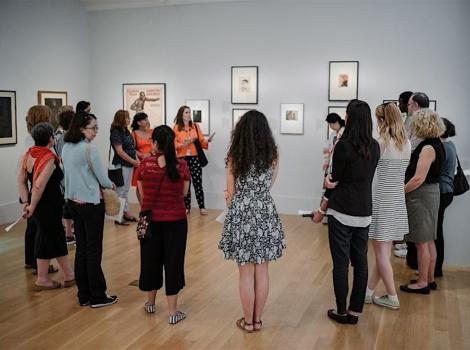 A group of people listen to a museum docent discuss a work of art hanging on a wall.